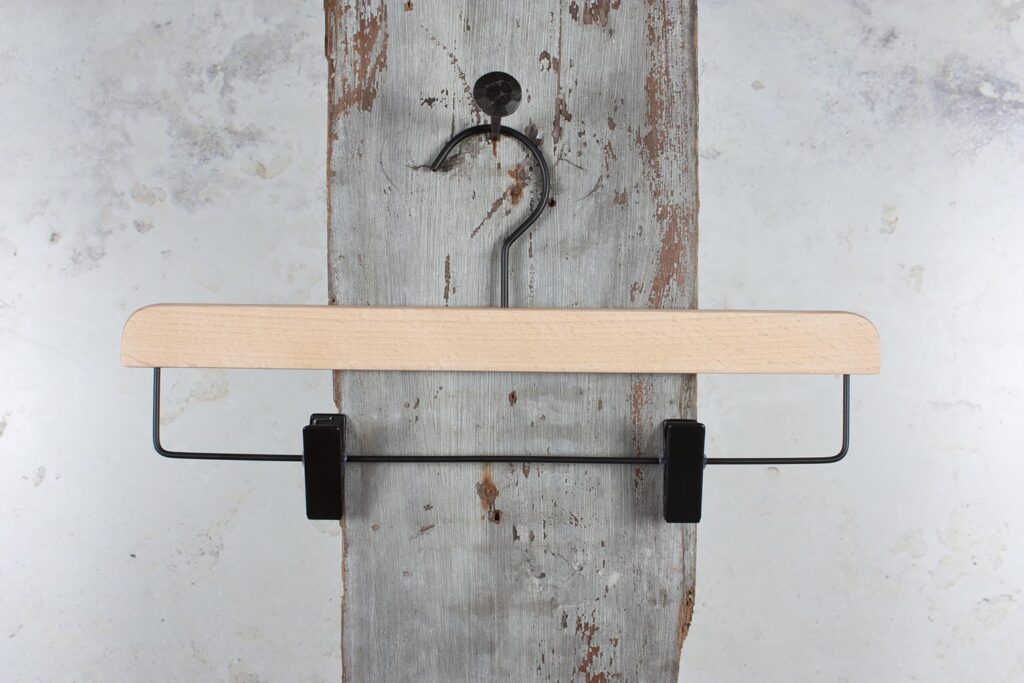 Hosenbügel aus Naturholz mit schwarzen Clips vor einer rustikalen Wand – klassisches Design für den Einzelhandel.